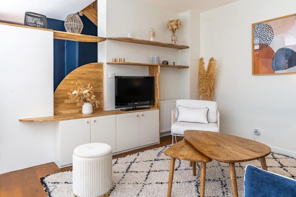 Salon avec meuble TV design sur mesure bois clair et blanc. Courbes élégantes, tapis berbère, pouf et tables basses gigognes.