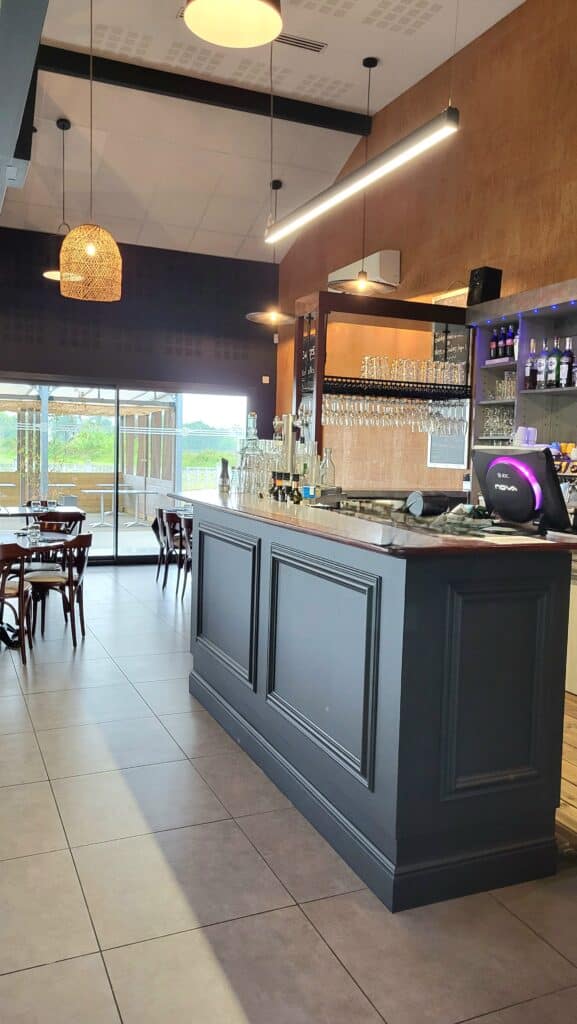 Intérieur de restaurant au design moderne. Bar gris foncé, verrerie et caisse JDC Nova. Tables en bois, vue sur la terrasse vitrée et la nature.