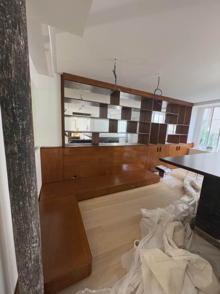 Meuble sur mesure intégré en bois verni (bibliothèque, banquette, rangements) avec étagères à miroirs. Pièce en rénovation avec poteau brut.