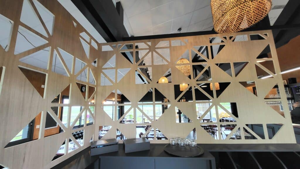 Claustra géométrique en bois clair divisant un espace de restauration. Comptoir sombre avec boîtes "GH" et luminaires en osier suspendus.