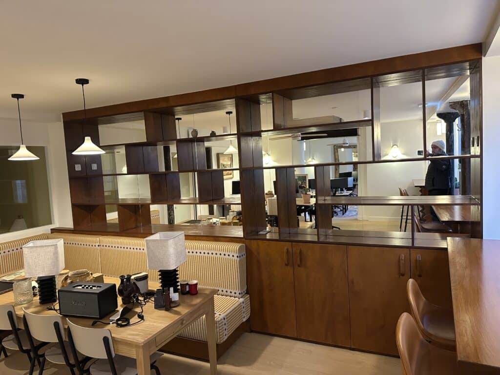 Design intérieur: Cloison bibliothèque en bois et miroirs séparant une salle à manger avec banquette rayée jaune d'un espace bureau moderne.
