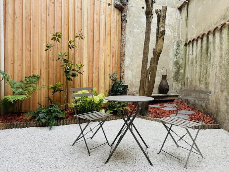 Cour intérieure zen avec table et deux chaises bistro sur gravier blanc. Clôture en bois, plantes vertes et vieux mur rustique à droite.