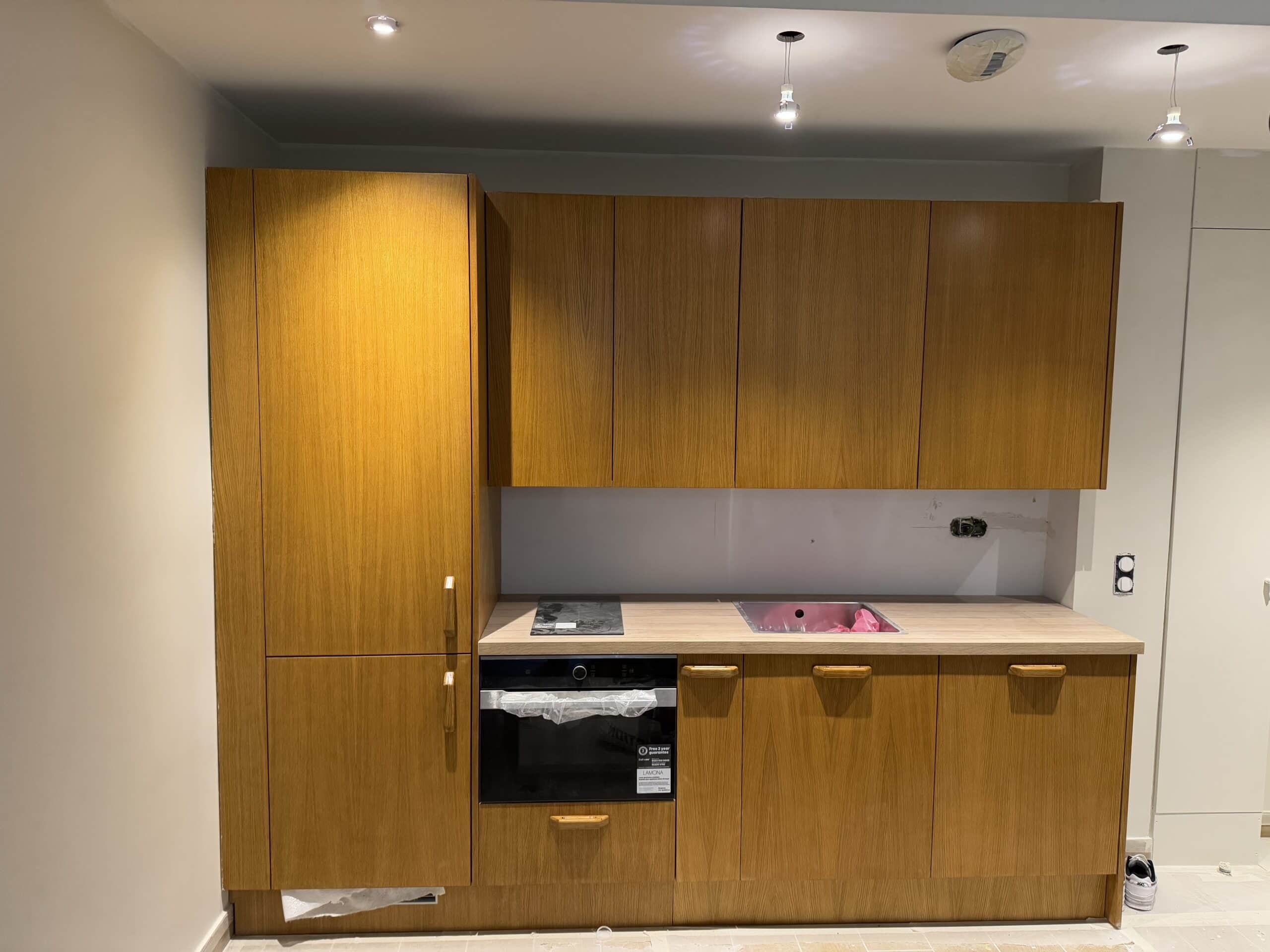 Installation de cuisine moderne : armoires en bois, comptoir clair. Four Lamona encastré, plaque de cuisson, et éclairage. Chantier.