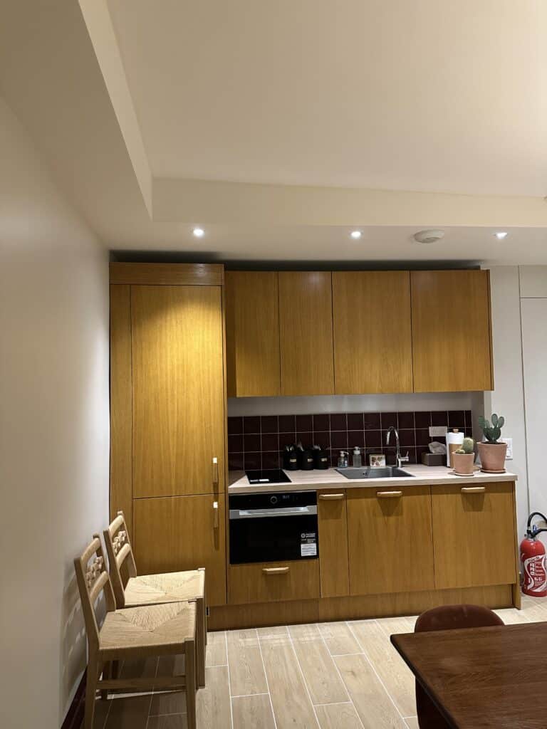 Cuisine moderne en bois clair (armoires hautes et basses) avec un four encastré Lamona et un dosseret de carreaux bordeaux. Deux chaises paille.