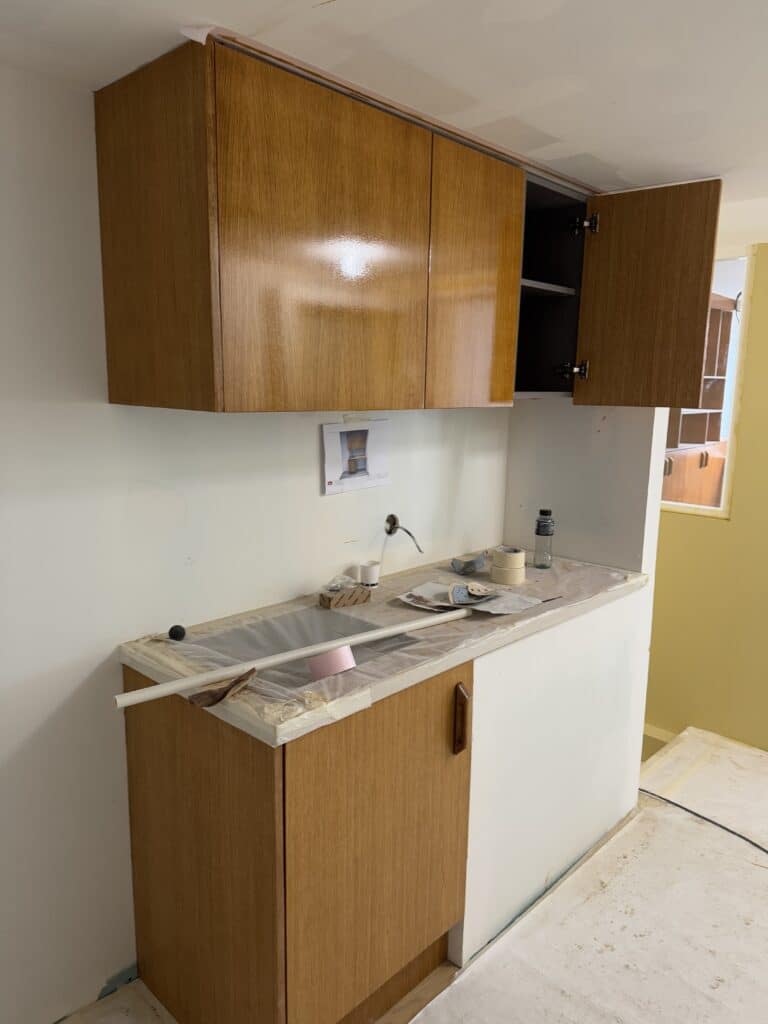 Cuisine en rénovation. Armoires murales et basses en bois clair installées. Plan de travail bâché, robinetterie absente. Chantier actif.