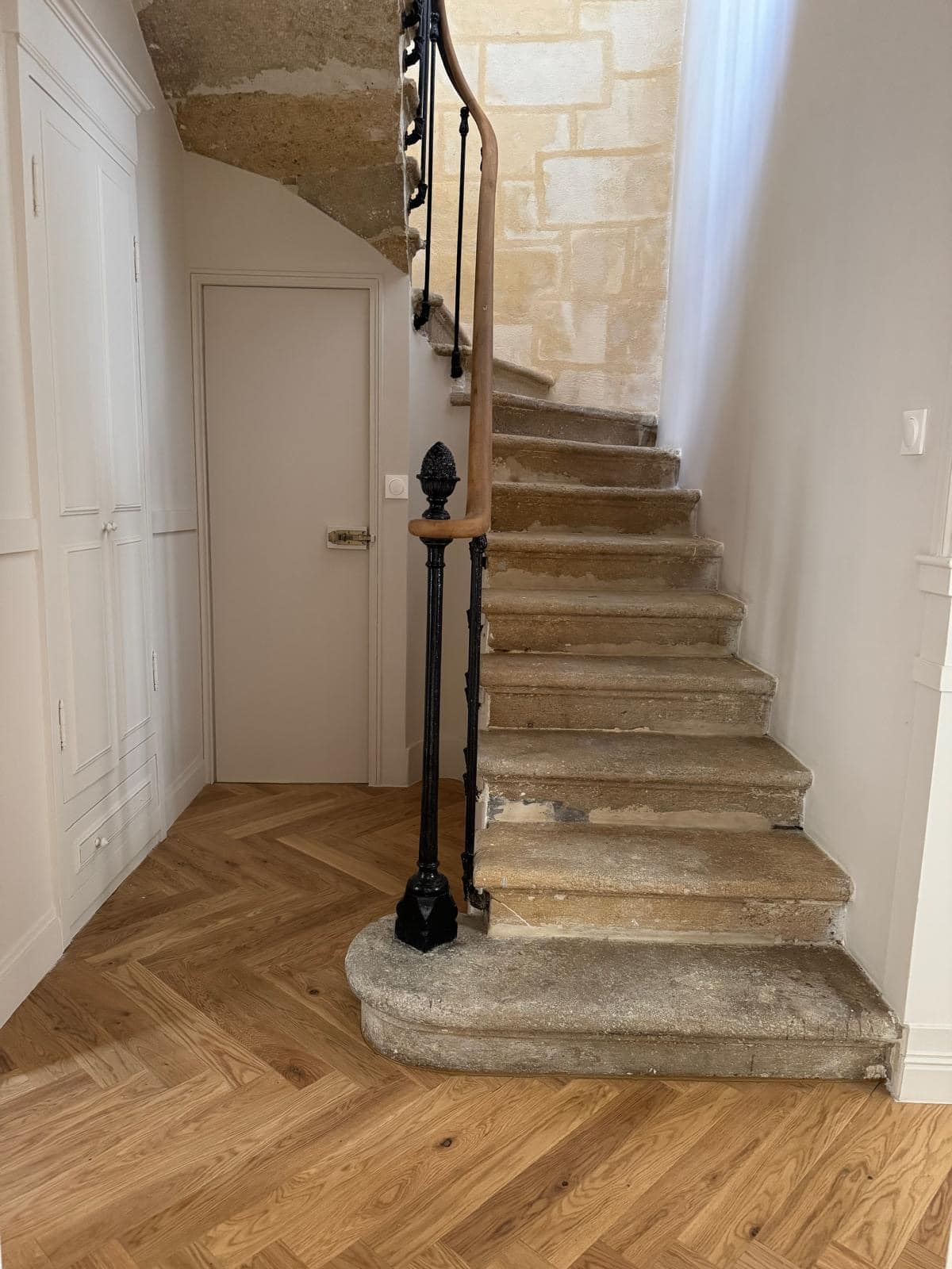 Escalier ancien en pierre brute avec rampe en fer forgé et main courante en bois, contrastant avec le parquet en chêne à chevrons.