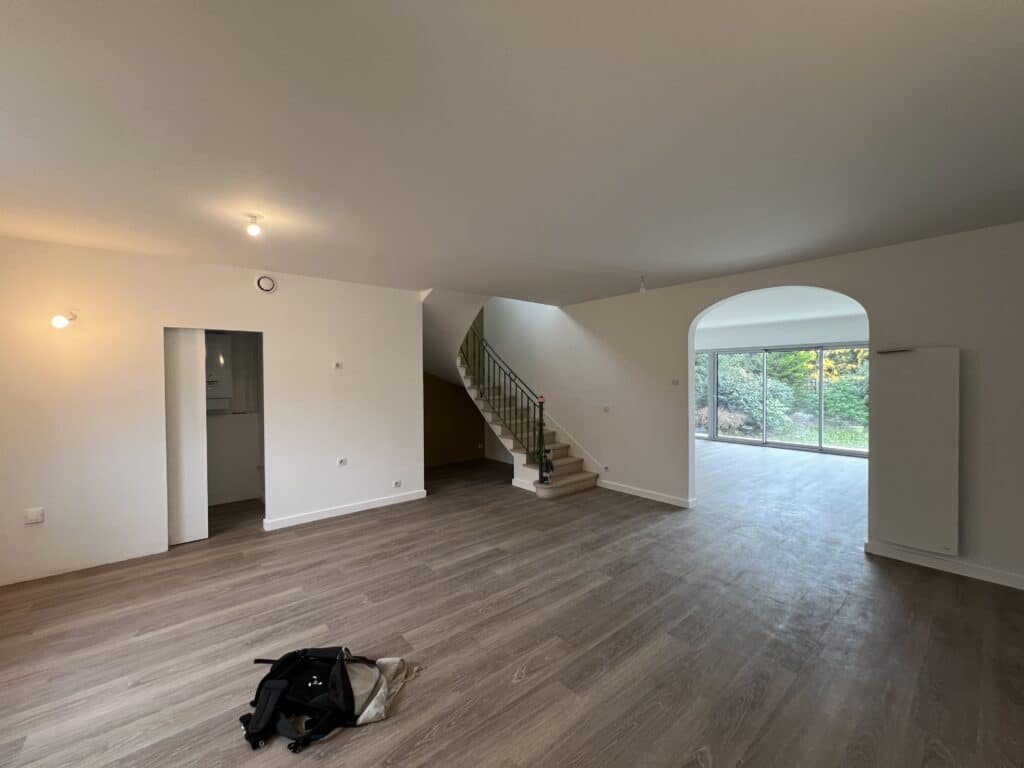 Salon en rénovation. Parquet moderne, escalier courbé avec rampe verte et ouverture en arche donnant sur le jardin. Murs blancs.