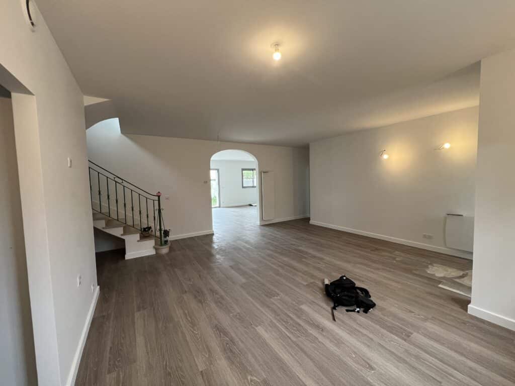 Intérieur moderne et lumineux d'un salon vide avec plancher flottant, escalier en fer forgé, murs blancs et arche vers la pièce suivante.