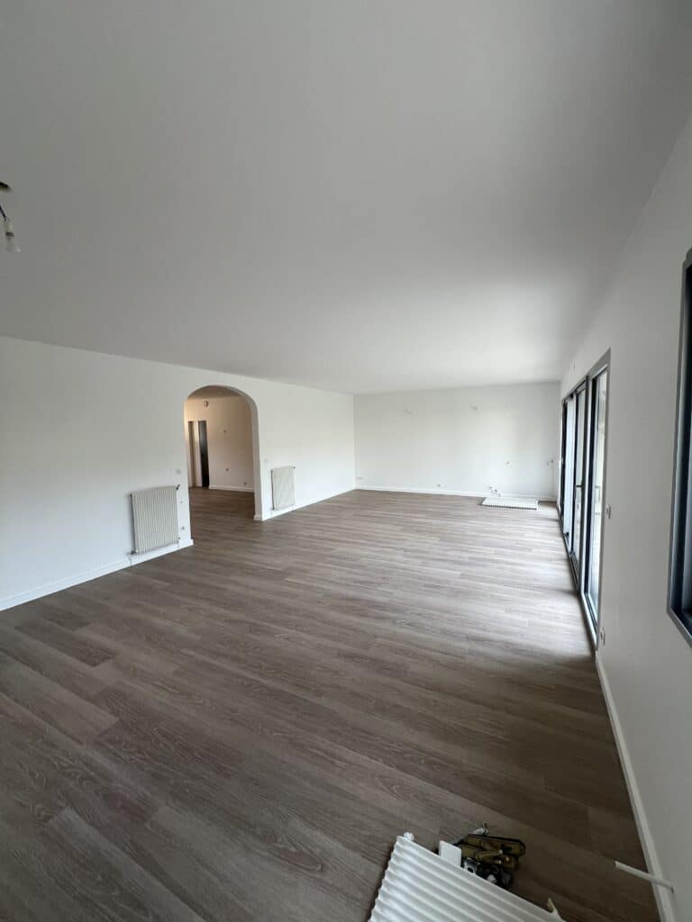 Grande pièce à vivre vide fraîchement rénovée. Sol stratifié brun, murs blancs, portes coulissantes vitrées et radiateurs.