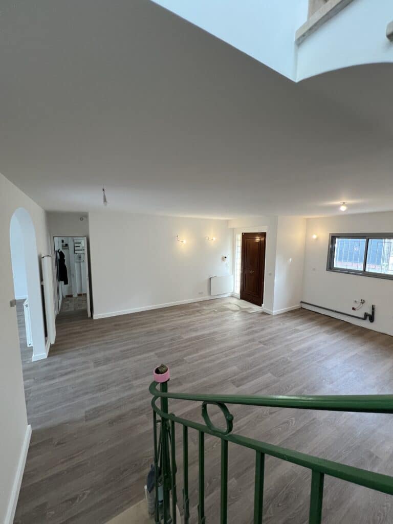 Grande pièce de vie vide après rénovation : parquet gris, murs blancs, escalier vert. Vue vers l'entrée et une petite salle d'eau.