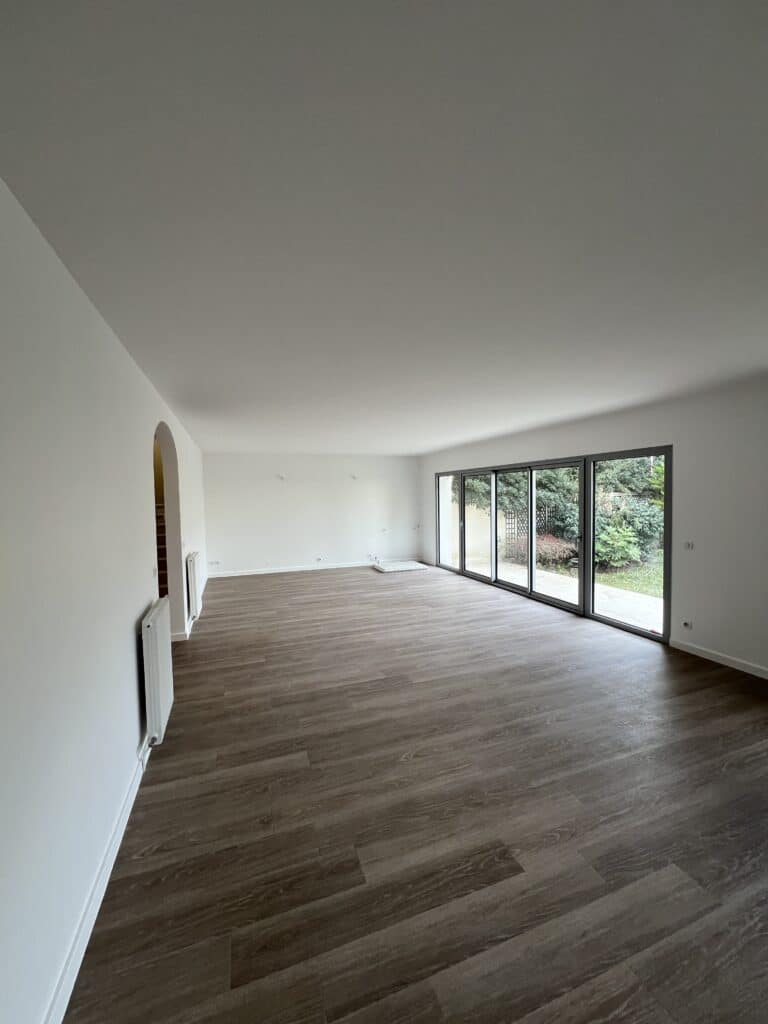 Salon vide et lumineux après rénovation. Murs blancs, parquet moderne et grande baie vitrée offrant vue sur le jardin.
