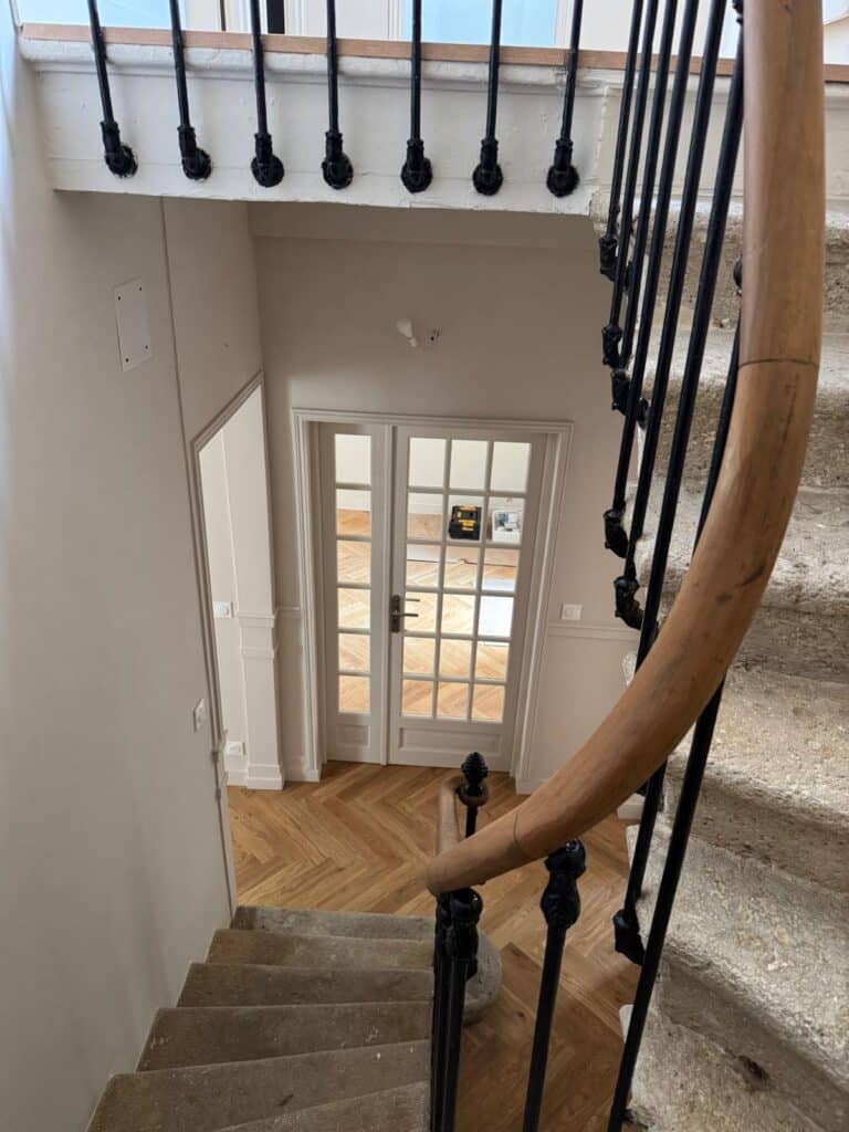 Vue plongeante sur un escalier en pierre et sa balustrade en fer forgé. Palier rénové avec parquet chevrons et doubles portes vitrées.