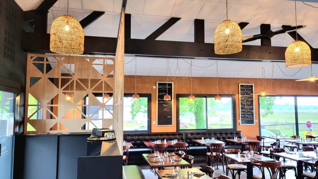 Restaurant moderne design. Claustra en bois, poutres noires. Grandes suspensions en rotin. Mobilier bistro, banquettes noires. Hélice d'avion au plafond. Vue sur l'aérodrome.