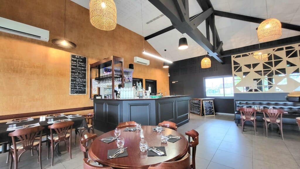 Salle de restaurant moderne avec bar central noir, poutres industrielles sombres et murs texturés ocre. Mobilier en bois, lumières tressées et menus à l'ardoise.