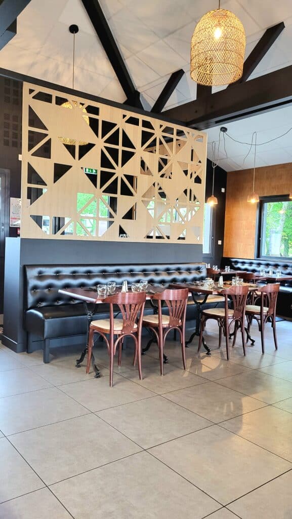 Restaurant moderne : banquettes noires, chaises bistrot bois foncé et tables dressées. Cloison décorative en bois clair à motifs géométriques.