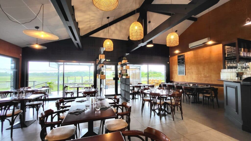 Intérieur de restaurant moderne avec poutres noires, murs ocre, chaises bistrot. Vue panoramique sur la pelouse. Menu "Cotelettes D'APIEAU" affiché.