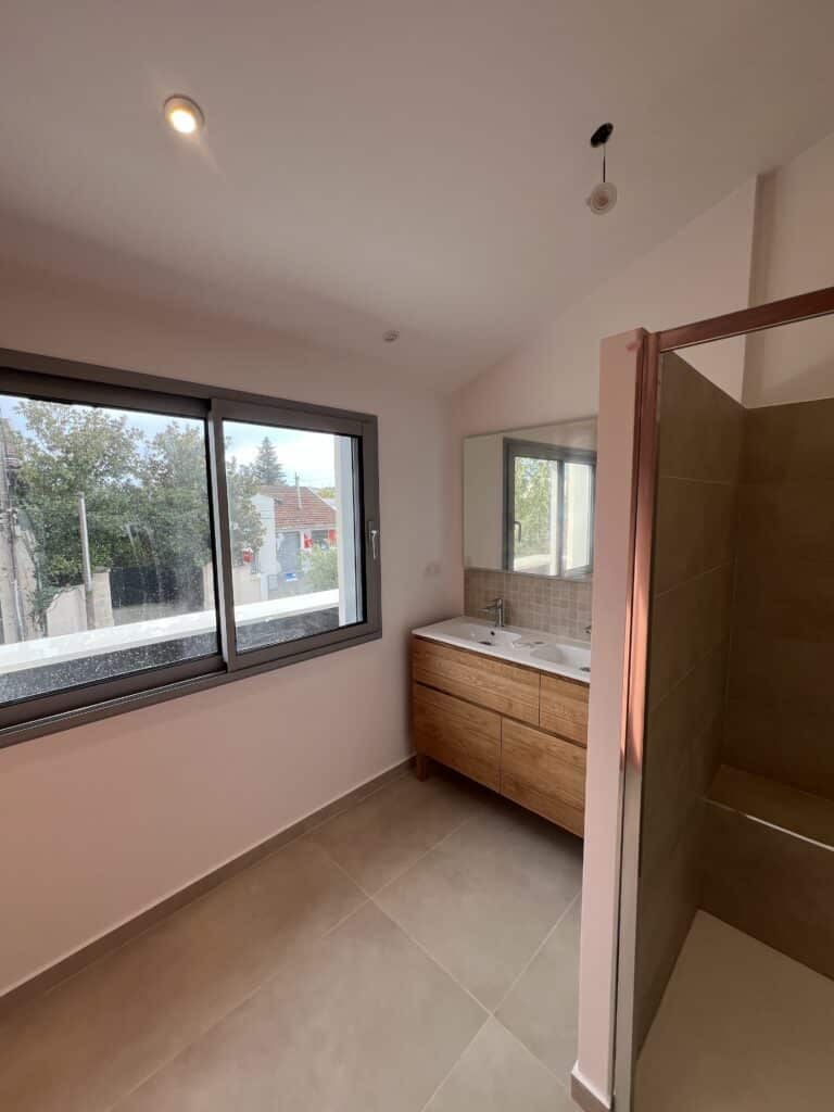 Salle de bain moderne en rénovation avec grande fenêtre, meuble double vasque en bois clair, murs roses et douche carrelée taupe.
