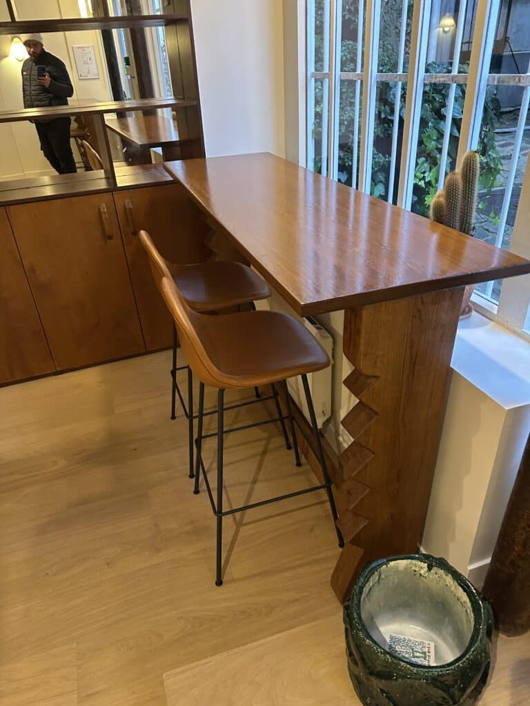 Longue table bar en bois verni sur base zigzag et deux tabourets hauts en cuir. Rangement mural. Reflet d'un homme au bonnet blanc dans le miroir.